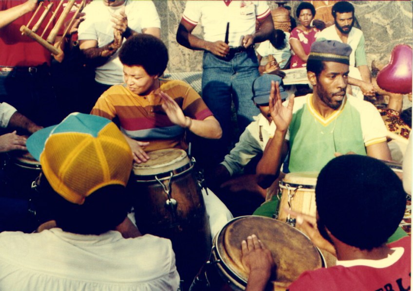 Terry Quaye and several African-American drummers playing congas in Washington Square, New York. Other people in background playing hand percussion.