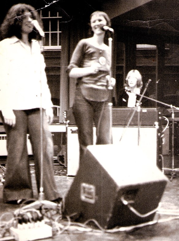 Black and white photograph of Vicky Scrivener (singing), MaggieNicols (singing), Josie Mitten (playing keyboards) performing. Vicky and Maggie smile at each other. 