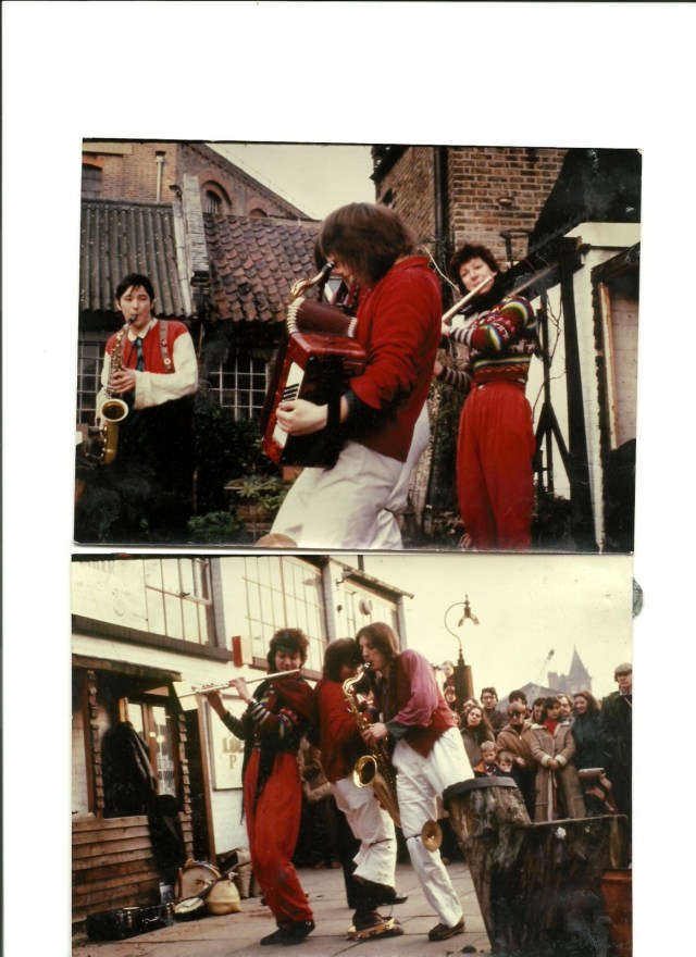 Colour photo of York Street Band playing outdoors, in a city square. Saxophone and flute players watch the accordionist. Image indicates that the band wore complementary red clothes when they performed. A small crowd is gathered to watch.