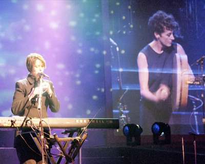 Live colour photograph of Zrazy performing, with bright blue stage lighting. Carole plays flute while standing behind electric keyboards and microphone, Maria at the front plays a bodhran, the Irish hand drum.