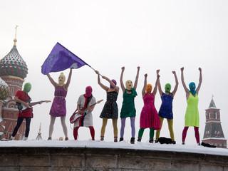 photo of eight Pussy Riot members on a rooftop, waving purple flag, fists in the air, two with guitars, wearing bright clothes and their trademark balaclavas, against a backdrop of Russian buildings