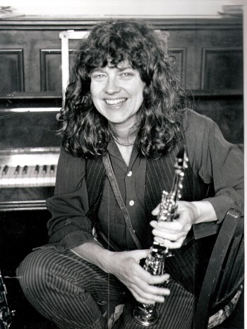 A portrait of Lindsay Cooper smiling, holding her sax, in front of a piano.