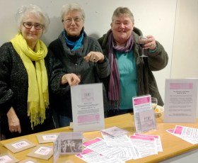 Three women smiling behind a stall displaying CDs, WLMA leaflets and posters, smiling and raising glass.